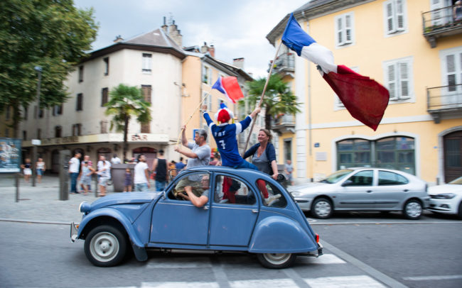 Des gens avec des drapeaux français sont dans une vieille 2CV dans les rues de Chambéry.
